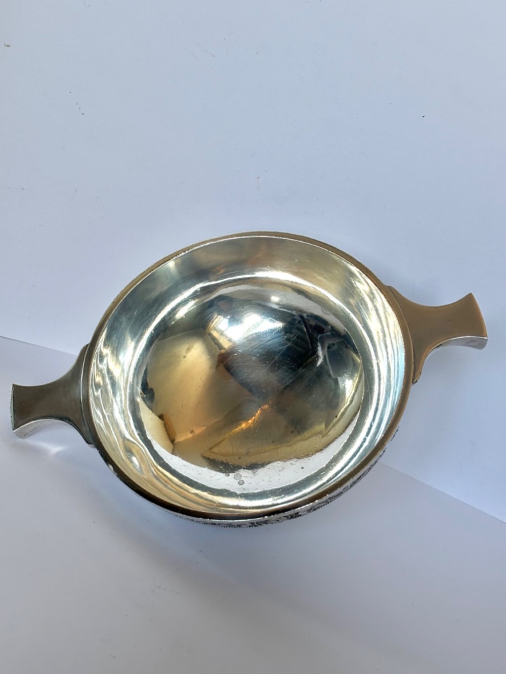 A shallow sterling silver quaich featuring two flattened lug handles, a circular pedestal base, and a band of embossed Celtic knotwork along the upper rim. View VI