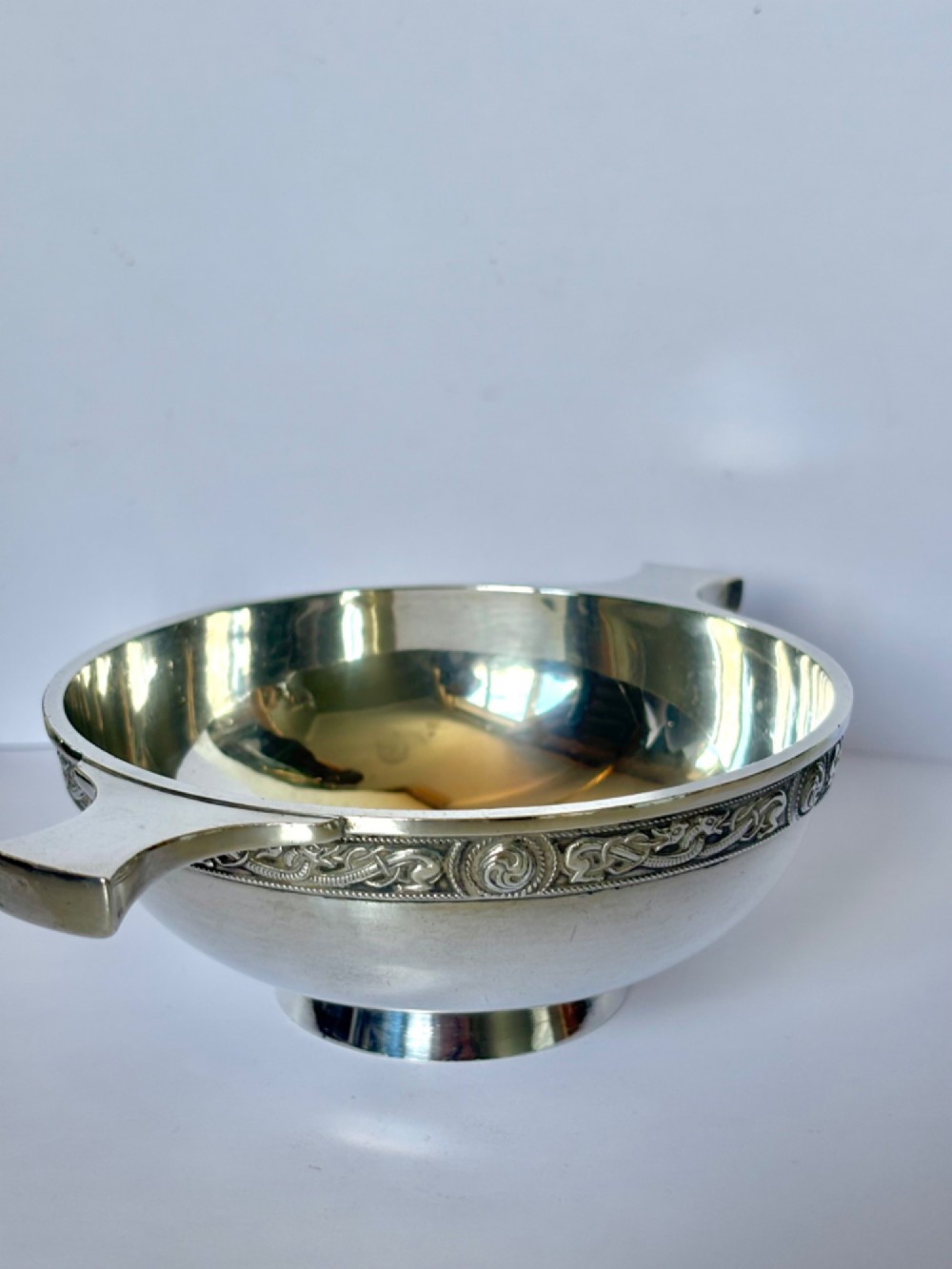 A shallow sterling silver quaich featuring two flattened lug handles, a circular pedestal base, and a band of embossed Celtic knotwork along the upper rim. View VII