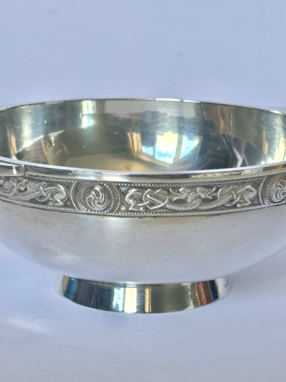 A shallow sterling silver quaich featuring two flattened lug handles, a circular pedestal base, and a band of embossed Celtic knotwork along the upper rim. View IX