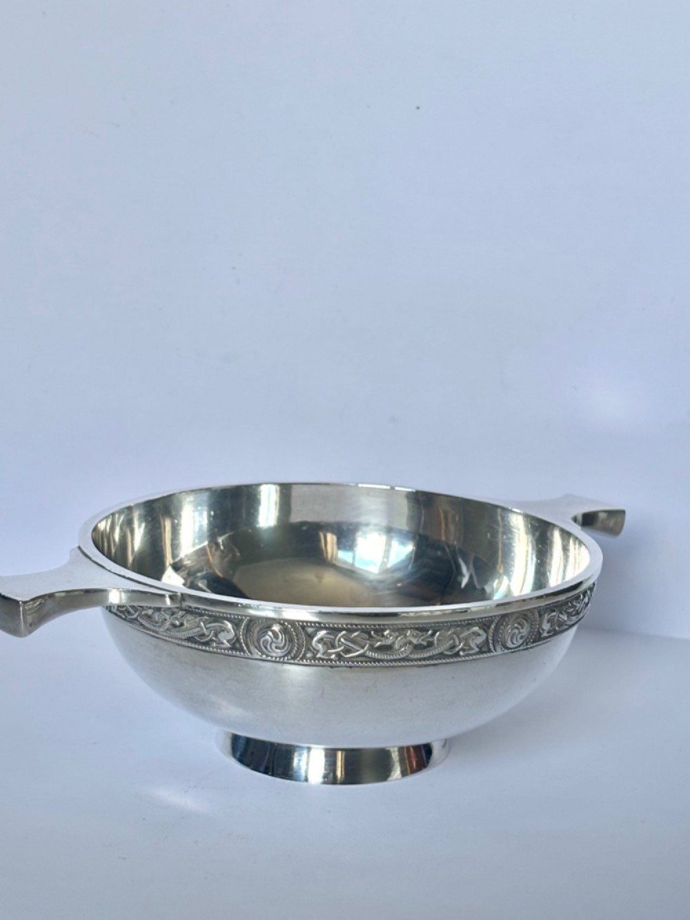 A shallow sterling silver quaich featuring two flattened lug handles, a circular pedestal base, and a band of embossed Celtic knotwork along the upper rim. View X