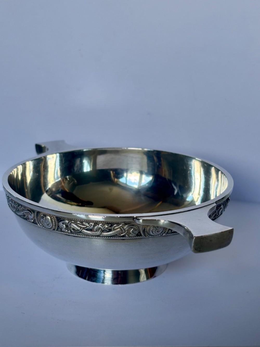A shallow sterling silver quaich featuring two flattened lug handles, a circular pedestal base, and a band of embossed Celtic knotwork along the upper rim. View XI