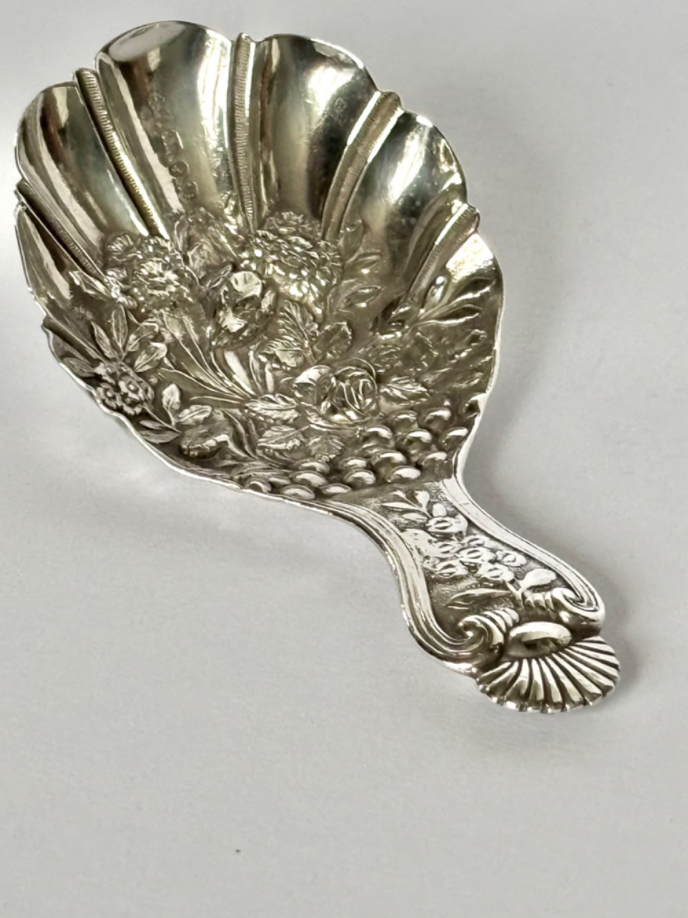 A Victorian sterling silver tea caddy spoon featuring a fluted shell-shaped bowl with embossed floral detailing and a hallmarked handle. View II
