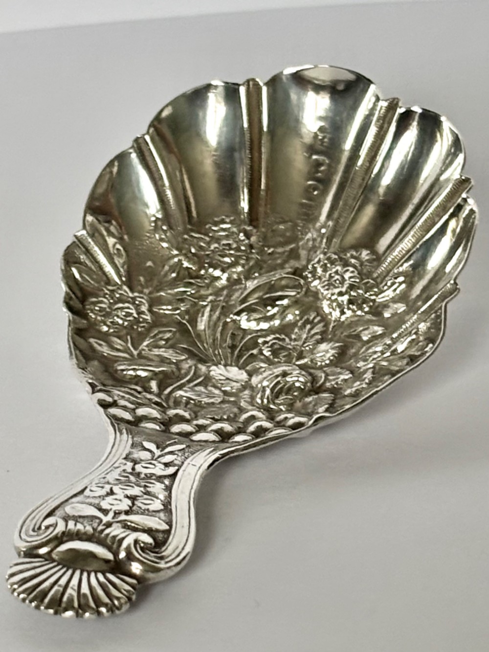 A Victorian sterling silver tea caddy spoon featuring a fluted shell-shaped bowl with embossed floral detailing and a hallmarked handle. View III