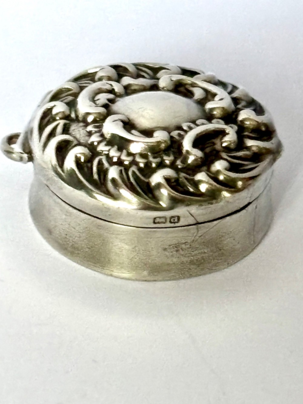 A small circular Edwardian sterling silver gilt trinket box with embossed scrollwork decoration on the lid. View V