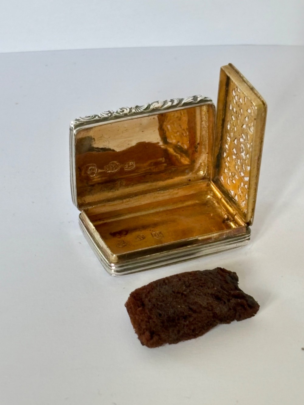 An open George IV rectangular silver-gilt vinaigrette showing internal hallmarks and a hinged pierced grille next to a small dark sponge.