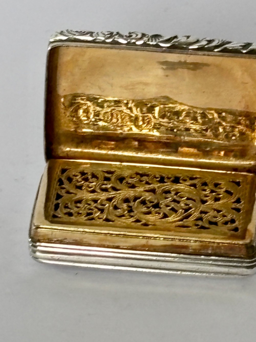 An open George IV rectangular silver-gilt vinaigrette showing internal hallmarks and a hinged pierced grille next to a small dark sponge. View IV