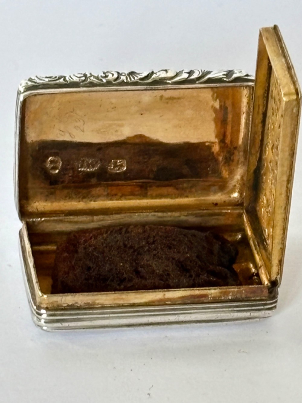 An open George IV rectangular silver-gilt vinaigrette showing internal hallmarks and a hinged pierced grille next to a small dark sponge. View VI