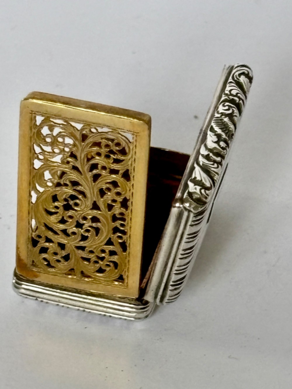 An open George IV rectangular silver-gilt vinaigrette showing internal hallmarks and a hinged pierced grille next to a small dark sponge. View XII