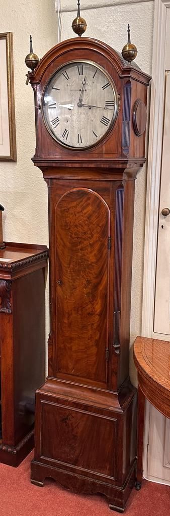 fig 1. An early nineteenth-century mahogany longcase clock featuring a silvered arched dial, brass finials, and flame mahogany veneers on the trunk door.
