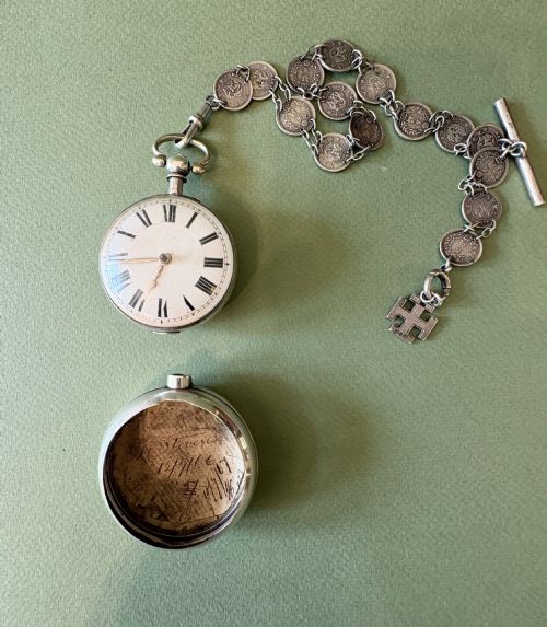 fig 1. An antique silver pair-cased pocket watch featuring a white enamel dial with black Roman numerals, connected to a silver chain formed of linked coins, a T-bar, and a small cross charm.