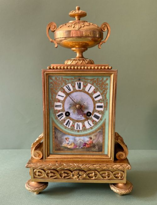 fig 1. A 19th-century French gilt metal mantel clock featuring hand-painted green porcelain panels and an urn finial, displayed inside a glass dome on a wooden base.