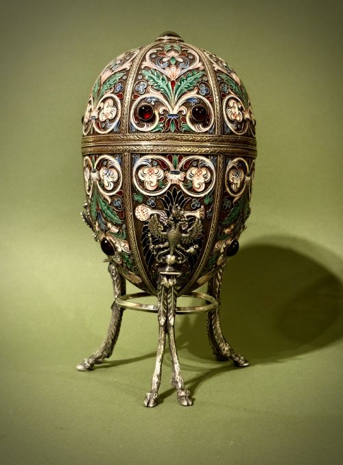 fig 1. A Russian silver egg decorated with polychrome floral enamel scrolls and set with cabochon stones, resting on a tripod silver stand with hoofed feet.