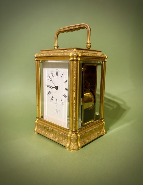 fig 2. An antique rectangular French carriage clock featuring a gilt brass case with engraved foliage, glass side panels, a top carrying handle, and a white enamel dial displaying Roman numerals.