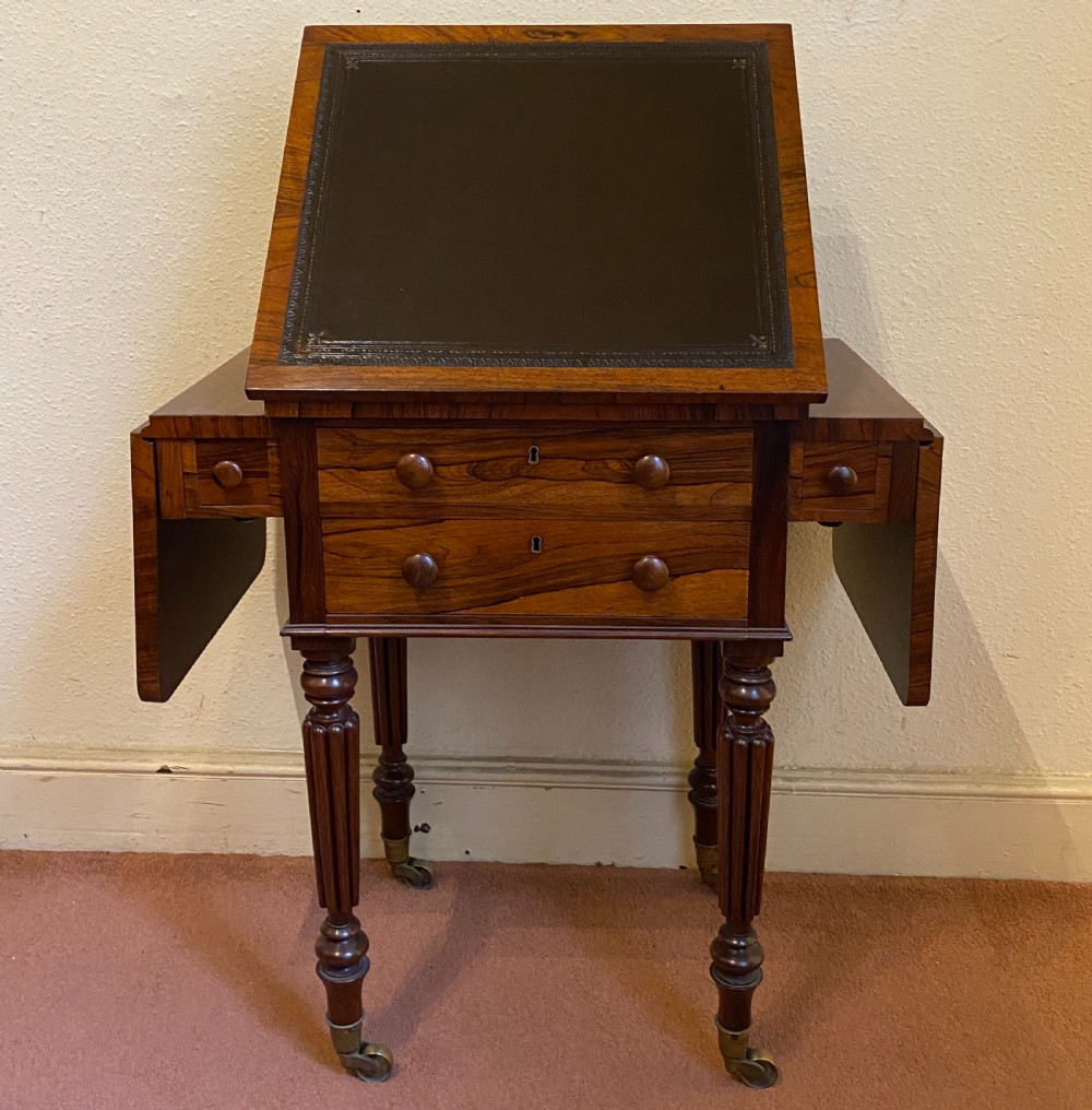 A William IV rosewood writing and work table featuring an adjustable leather-inset writing slope, drop-leaf sides, two drawers, and reeded turned legs on brass castors.