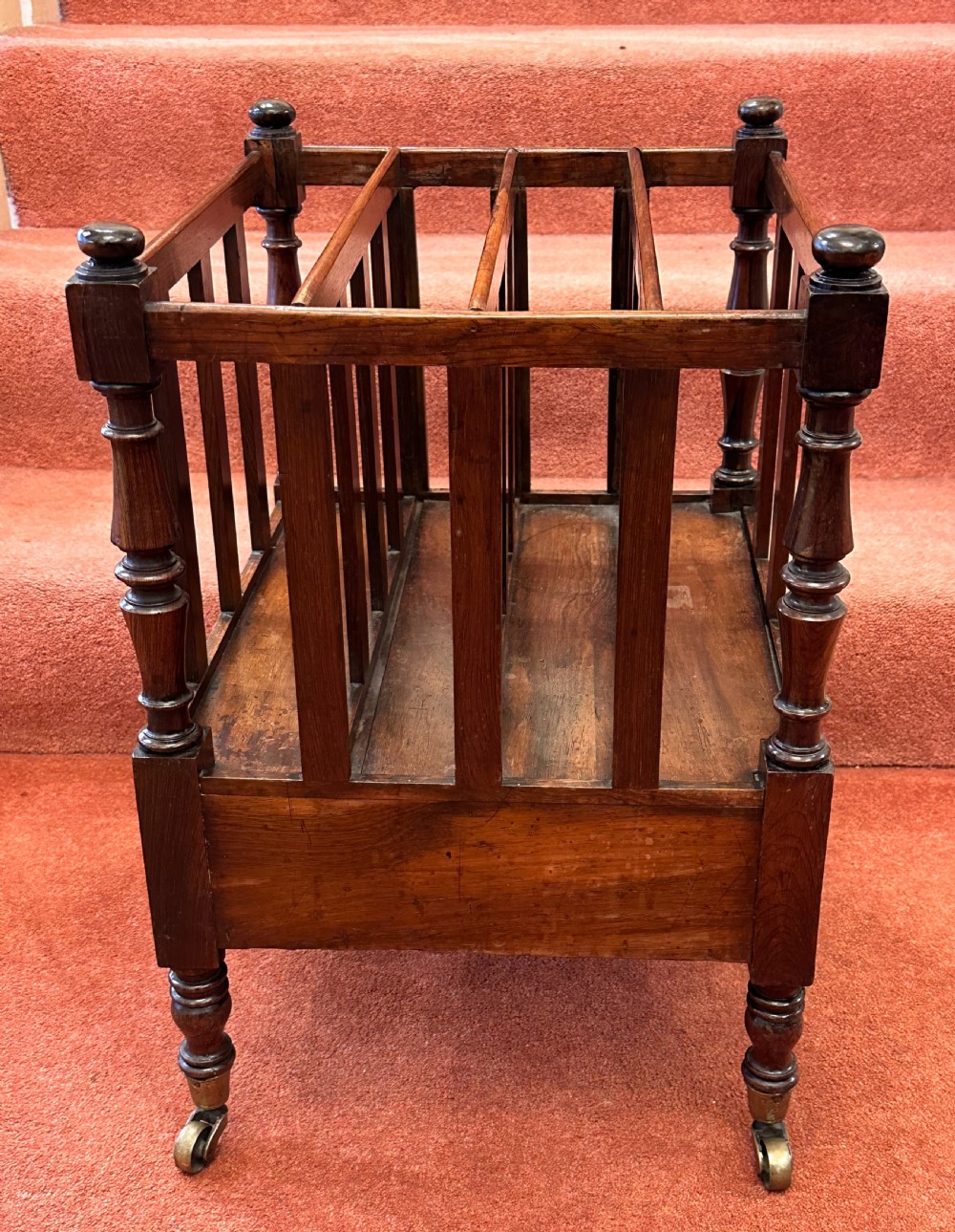 A 19th-century mahogany music Canterbury featuring four divided compartments, turned corner posts with finials, a single lower drawer with wooden knobs, and turned legs on brass castors. View VI