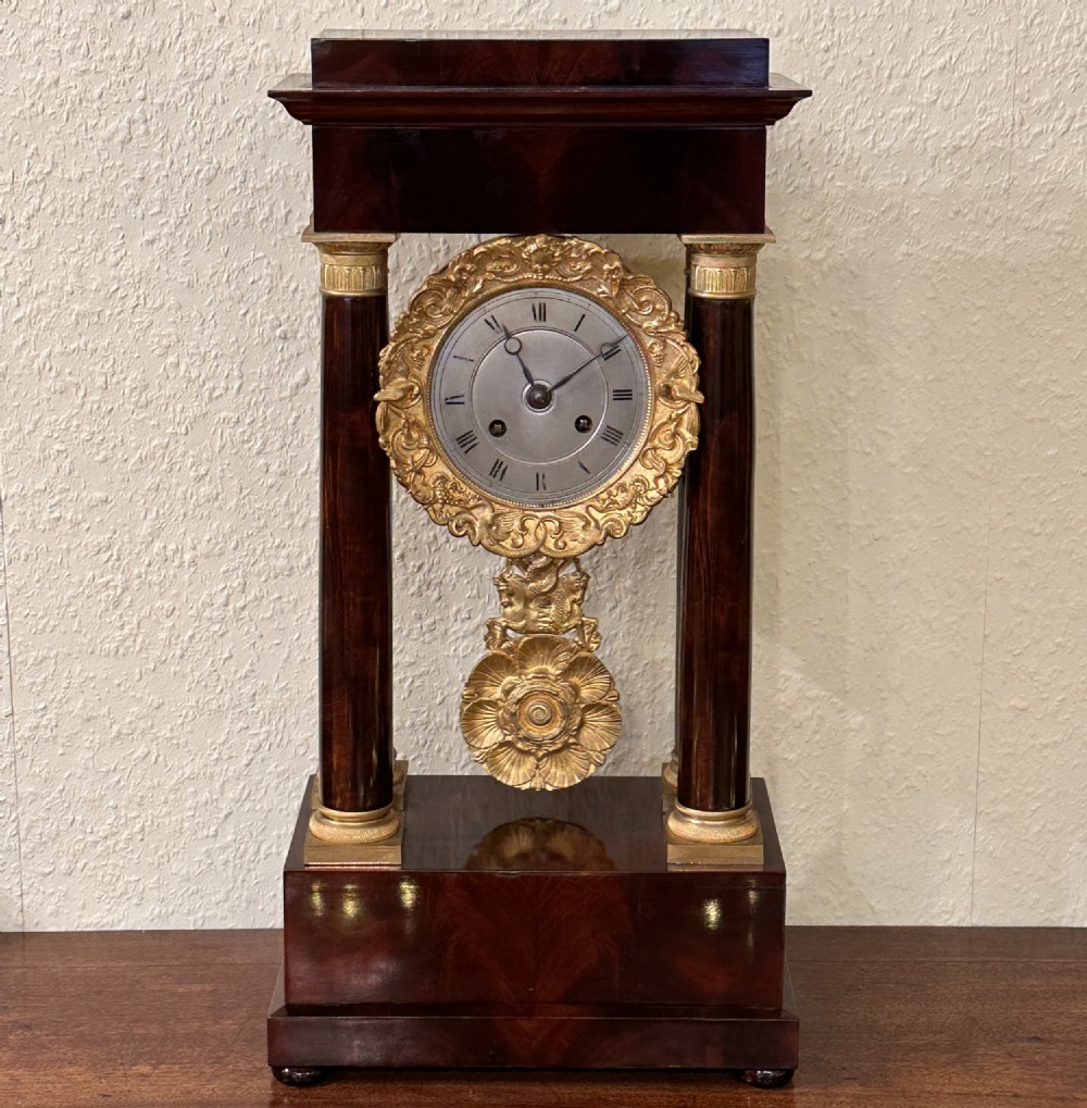A 19th-century mahogany portico clock featuring architectural columns, a gilt-bronze floral bezel, a silvered dial with Roman numerals, and an ornate swinging pendulum.