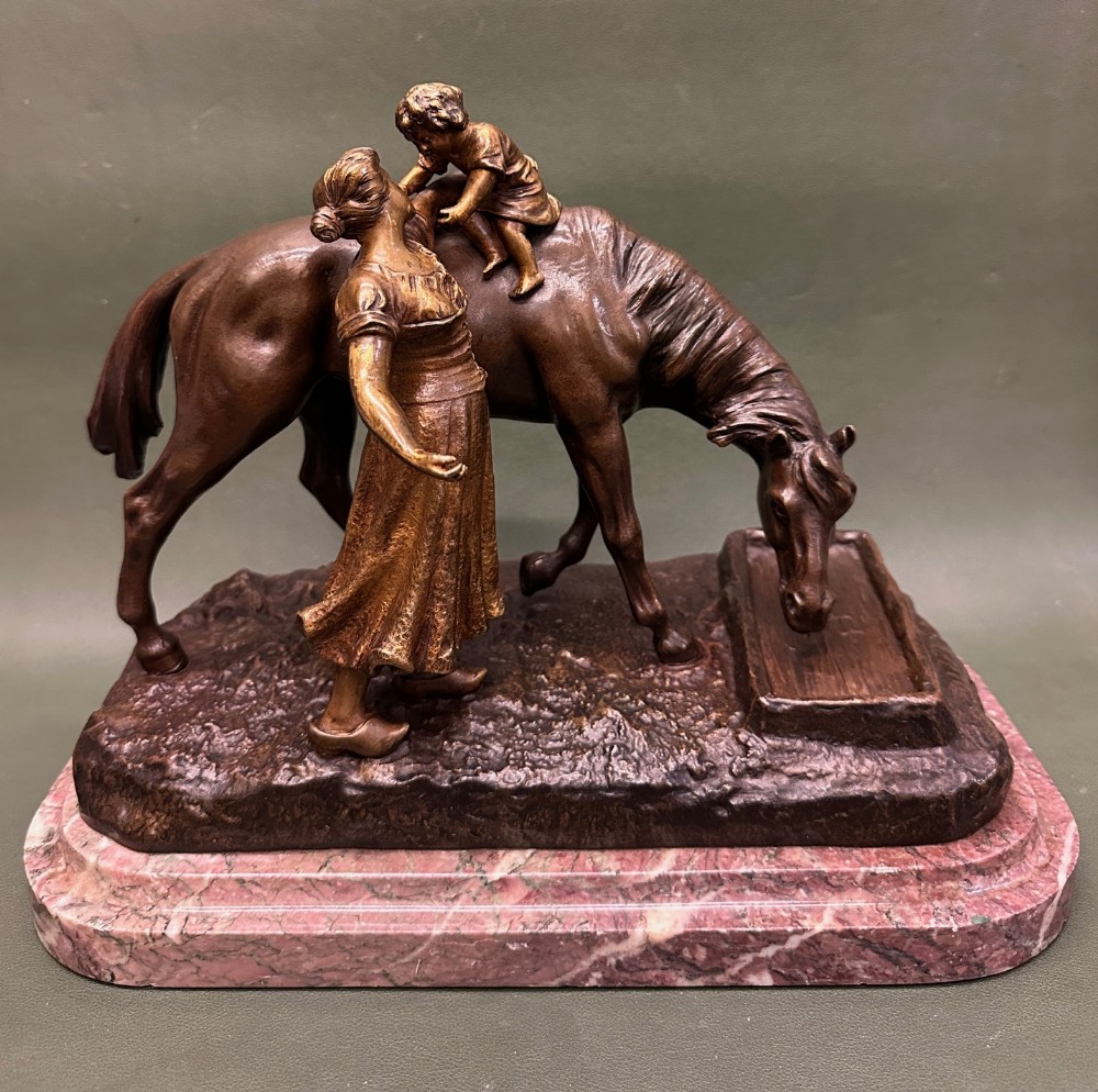 A patinated bronze sculpture depicting a mother and child standing beside a horse drinking from a trough, mounted on a pink veined marble base.