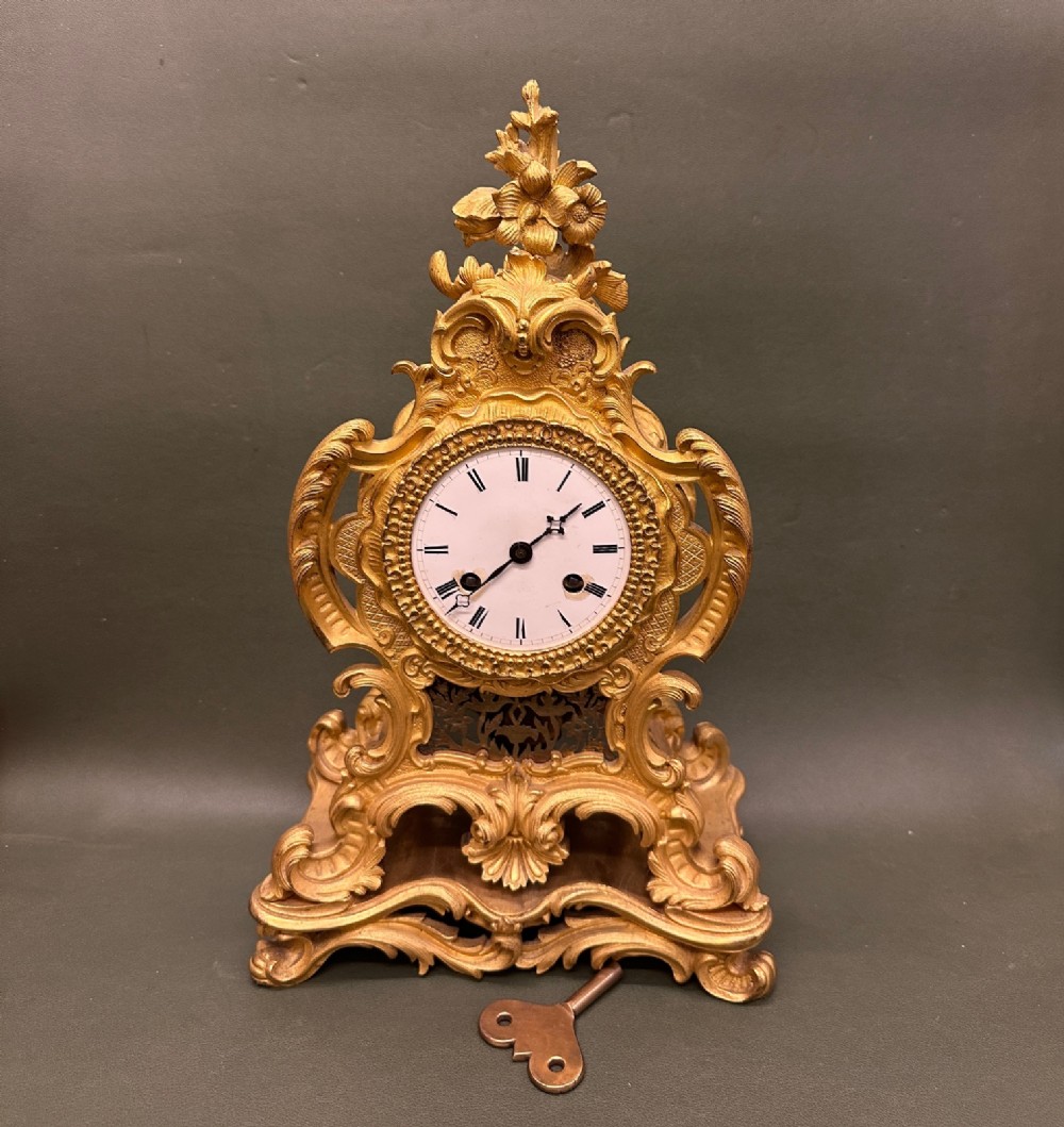 A 19th-century French Rococo revival gilded mantel clock featuring intricate scrollwork, a floral finial, a white enamel dial with Roman numerals, and a separate winding key.