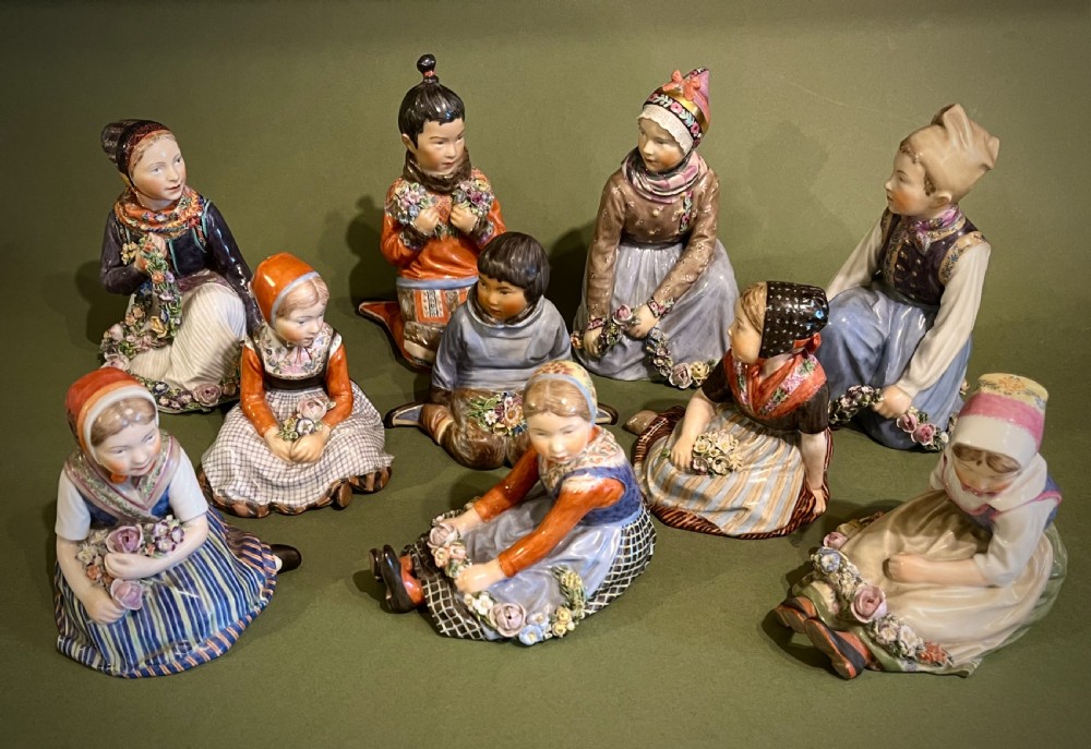 A set of ten hand-painted Royal Copenhagen porcelain figurines depicting seated children wearing traditional Danish regional costumes and floral garlands.