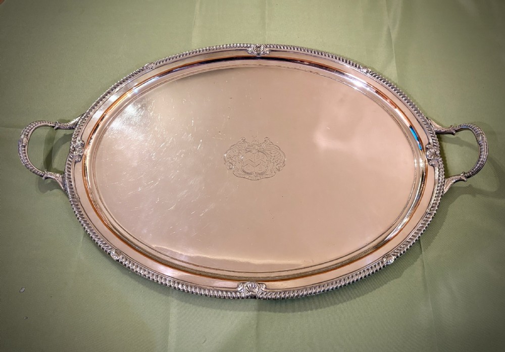 An oval Old Sheffield plate serving tray featuring two handles, a gadrooned rim with shell motifs, and an engraved armorial crest in the centre.