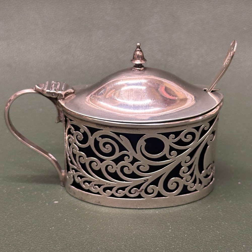 An oval pierced silver mustard pot with a domed lid, scroll handle, and visible blue glass liner, accompanied by a small spoon.