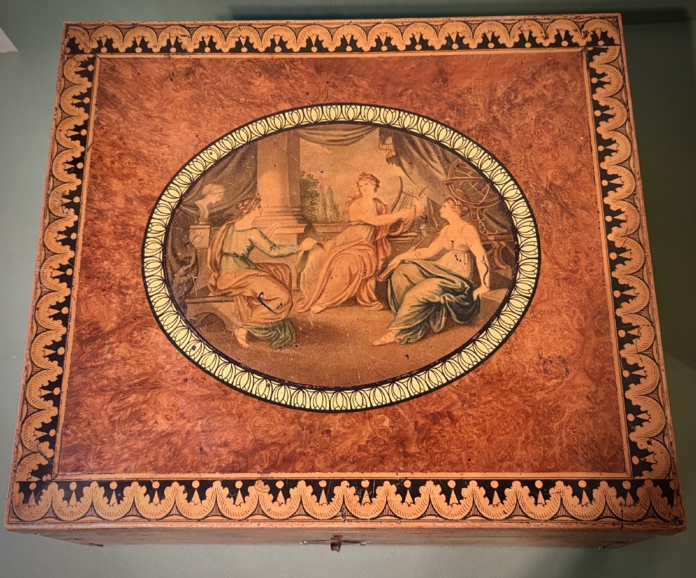 A square Regency burr walnut workbox with a central classical medallion print, decorative printed border, brass side handles, and a lower drawer. View VI