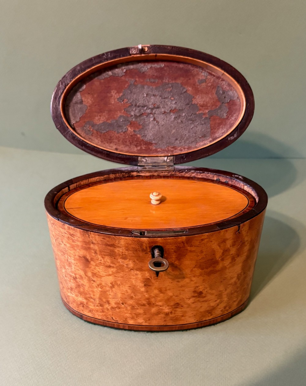 An oval George III satinwood tea caddy featuring inlaid crossbanding, a dark keyhole escutcheon, and an accompanying metal key. View IV