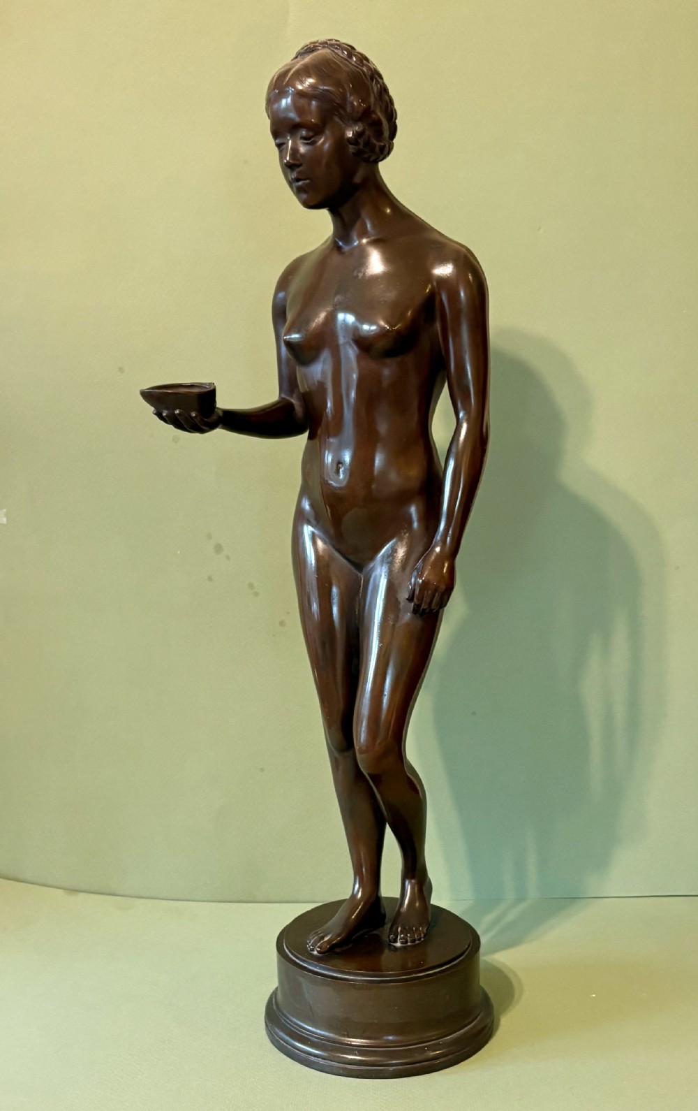 A dark patinated bronze sculpture depicting a standing female nude holding a small vessel, resting on a rounded stepped base.