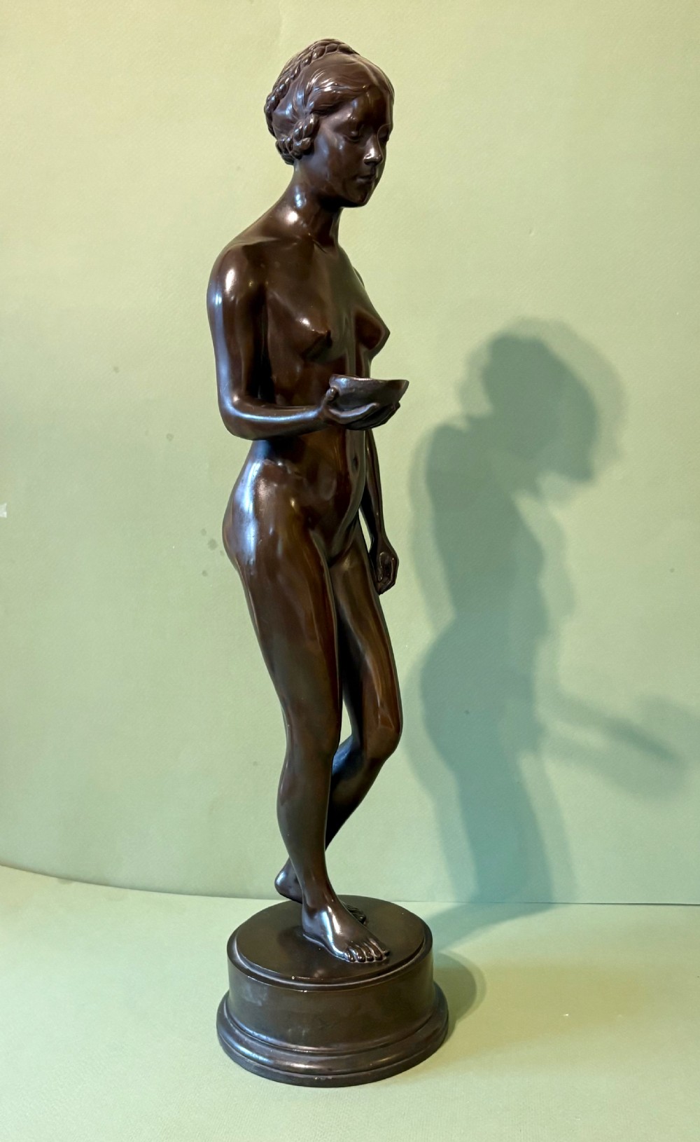A dark patinated bronze sculpture depicting a standing female nude holding a small vessel, resting on a rounded stepped base. View III