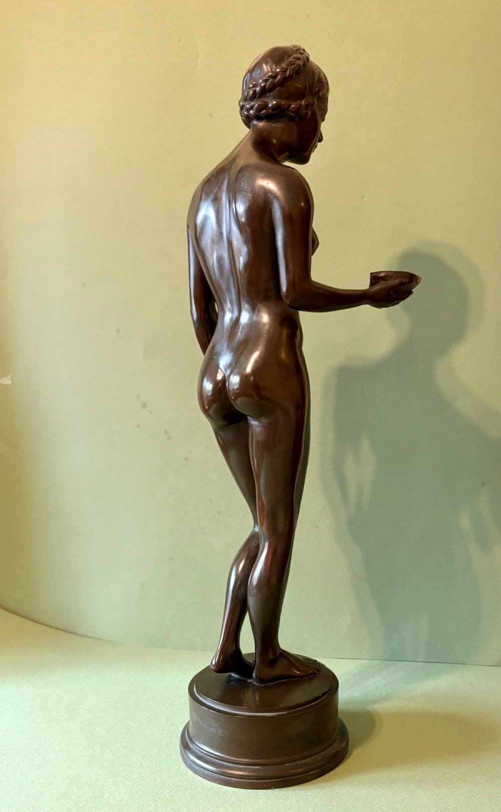 A dark patinated bronze sculpture depicting a standing female nude holding a small vessel, resting on a rounded stepped base. View IV