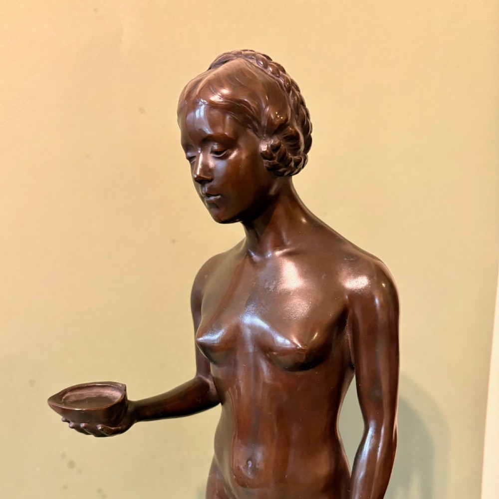 A dark patinated bronze sculpture depicting a standing female nude holding a small vessel, resting on a rounded stepped base. View V