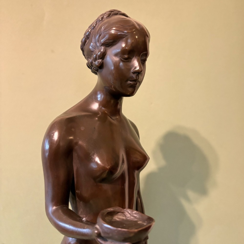 A dark patinated bronze sculpture depicting a standing female nude holding a small vessel, resting on a rounded stepped base. View VI