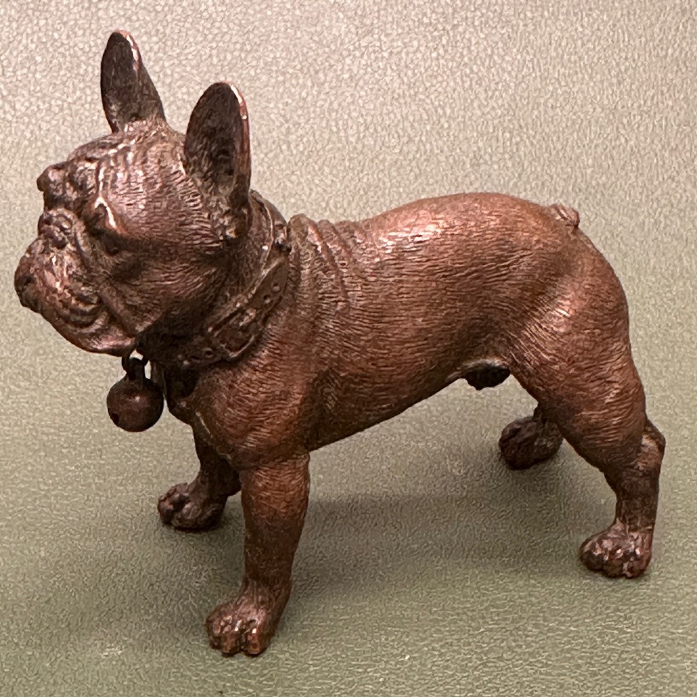 A cast bronze sculpture of a French bulldog standing upright with a buckled collar and hanging bell.