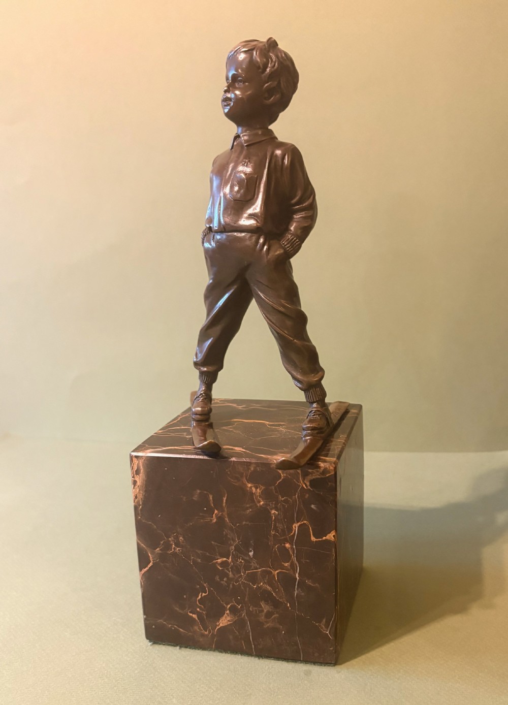 A dark bronze figurine depicting a young boy on skis mounted on a heavy, reddish-brown veined marble plinth.