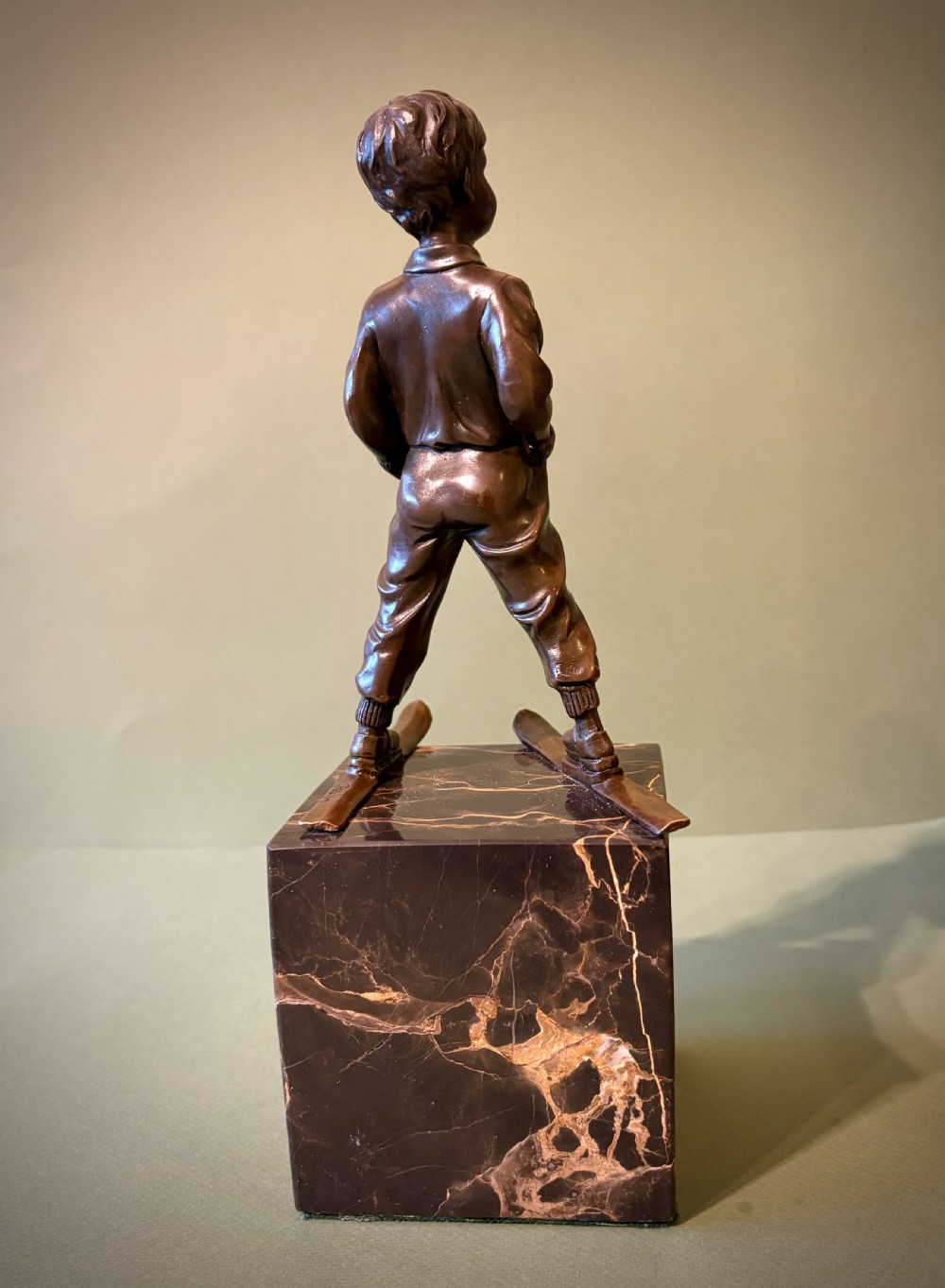 A dark bronze figurine depicting a young boy on skis mounted on a heavy, reddish-brown veined marble plinth. View III