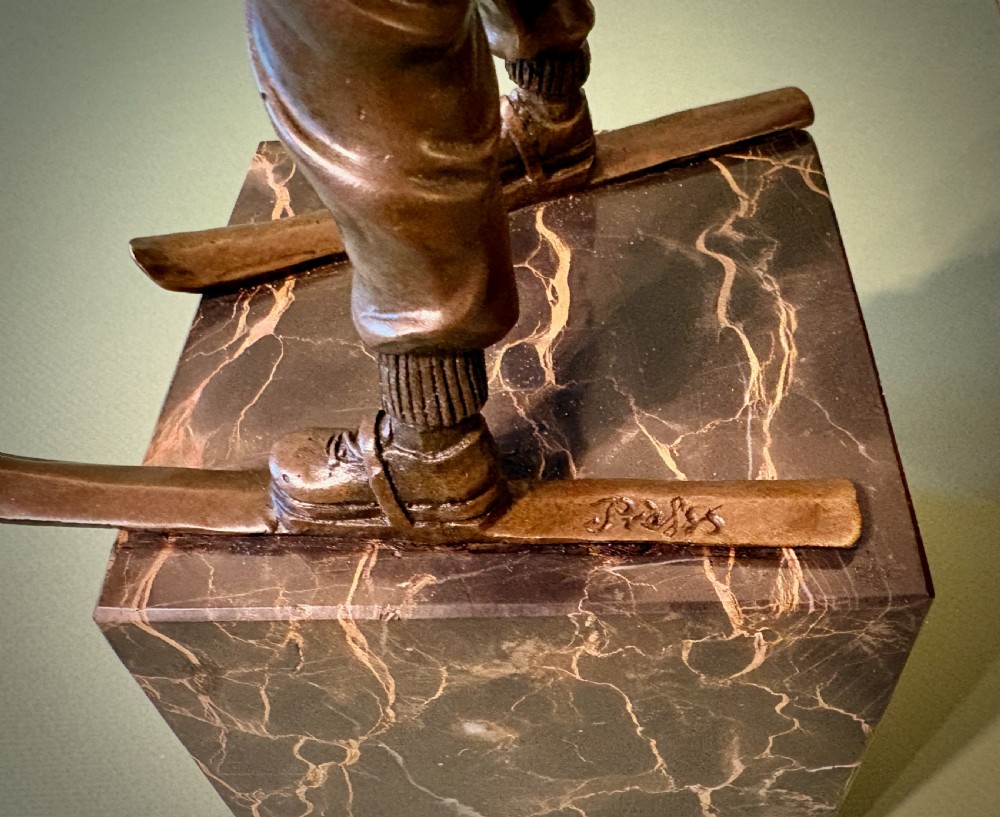 A dark bronze figurine depicting a young boy on skis mounted on a heavy, reddish-brown veined marble plinth. View IV