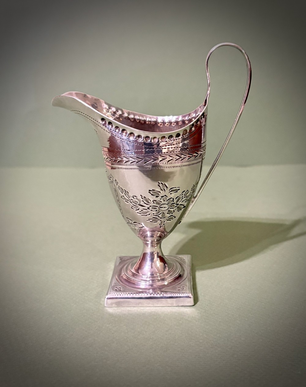 A George III sterling silver helmet-shaped cream jug featuring bright-cut floral engraving, a high loop handle, and a square pedestal base.