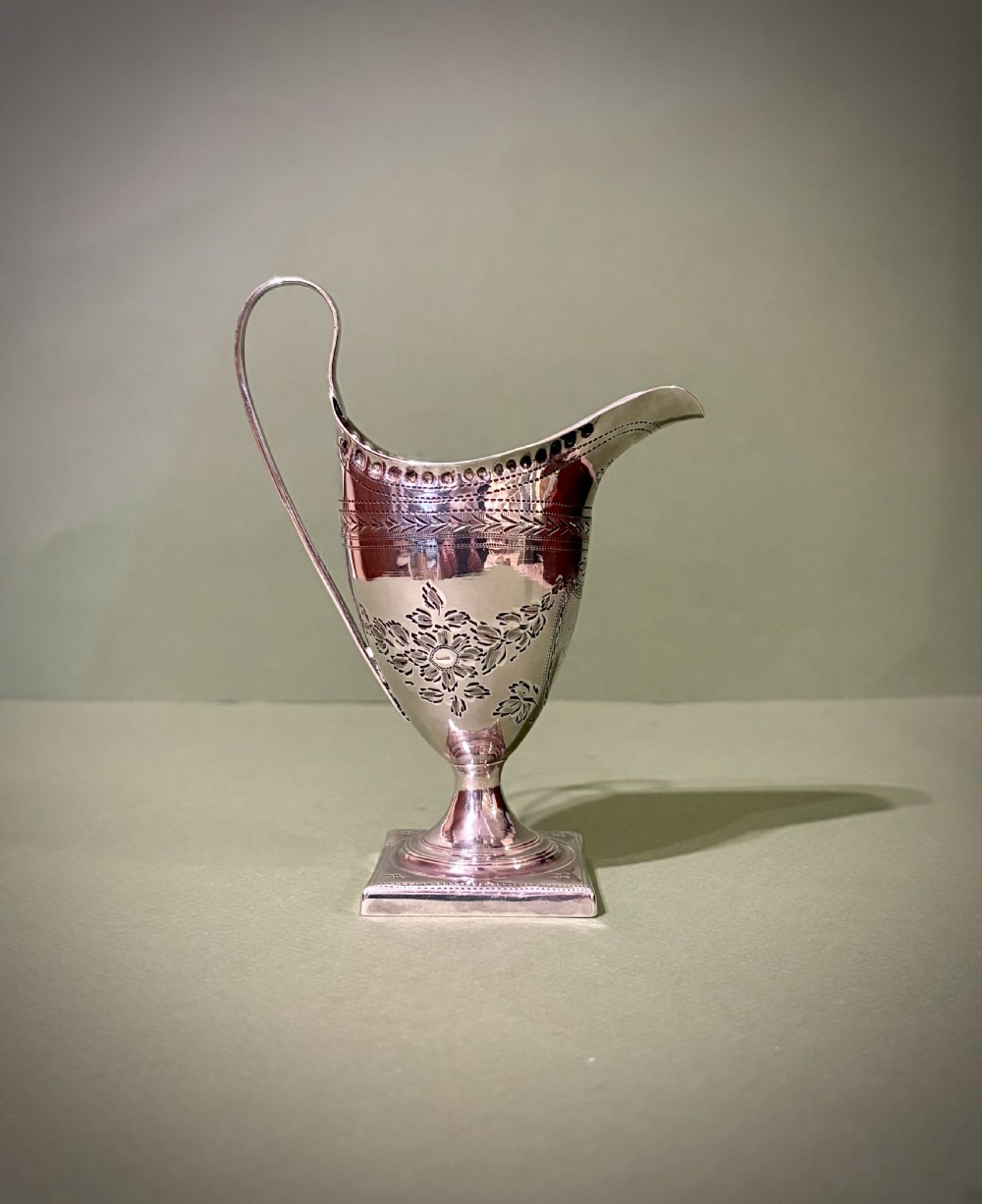 A George III sterling silver helmet-shaped cream jug featuring bright-cut floral engraving, a high loop handle, and a square pedestal base. View II