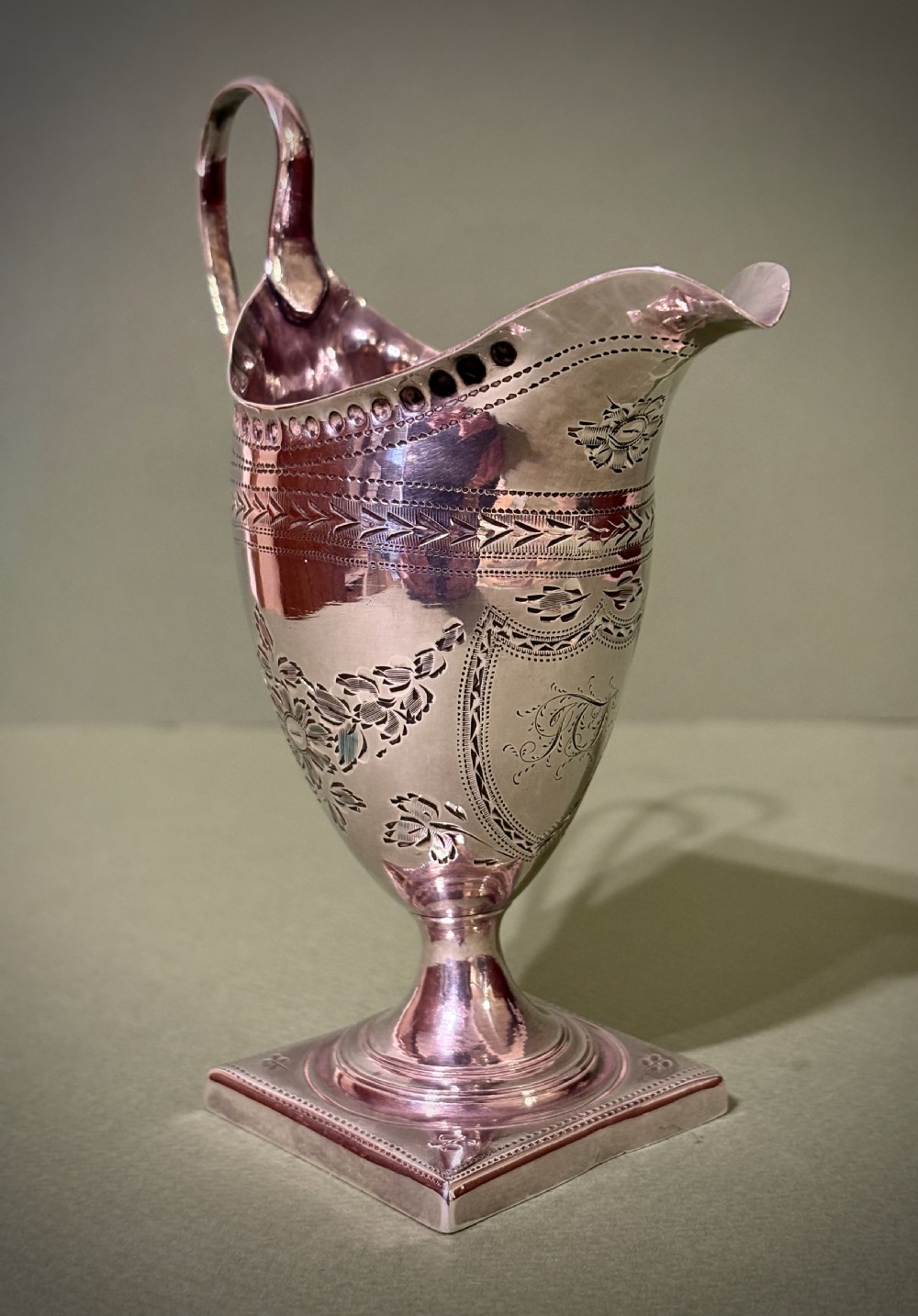 A George III sterling silver helmet-shaped cream jug featuring bright-cut floral engraving, a high loop handle, and a square pedestal base. View III