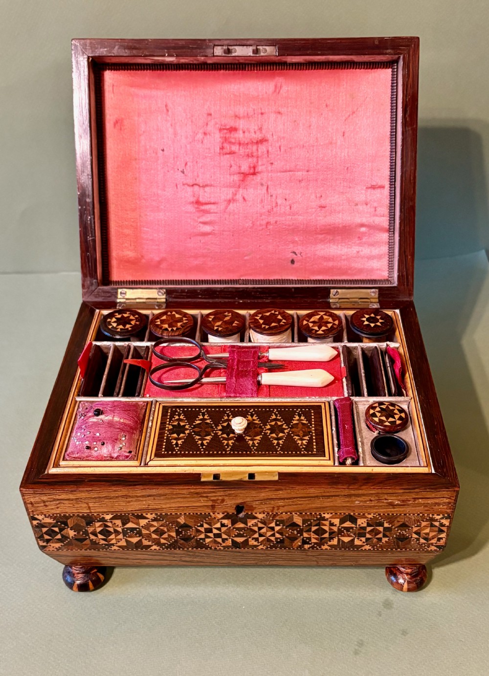 An open Victorian rosewood sewing box featuring geometric Tunbridge ware mosaic inlay, a pink silk interior, and fitted compartments containing sewing tools.