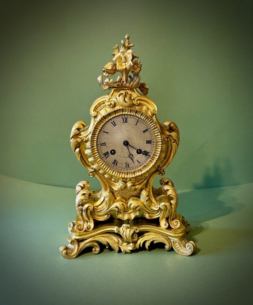 An ornate French Rococo style ormolu mantel clock featuring elaborate leaf scrollwork, a floral finial, and a circular dial with Roman numerals.