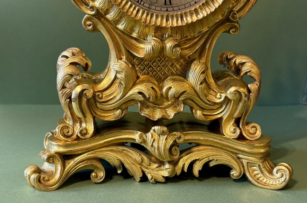 An ornate French Rococo style ormolu mantel clock featuring elaborate leaf scrollwork, a floral finial, and a circular dial with Roman numerals. View II