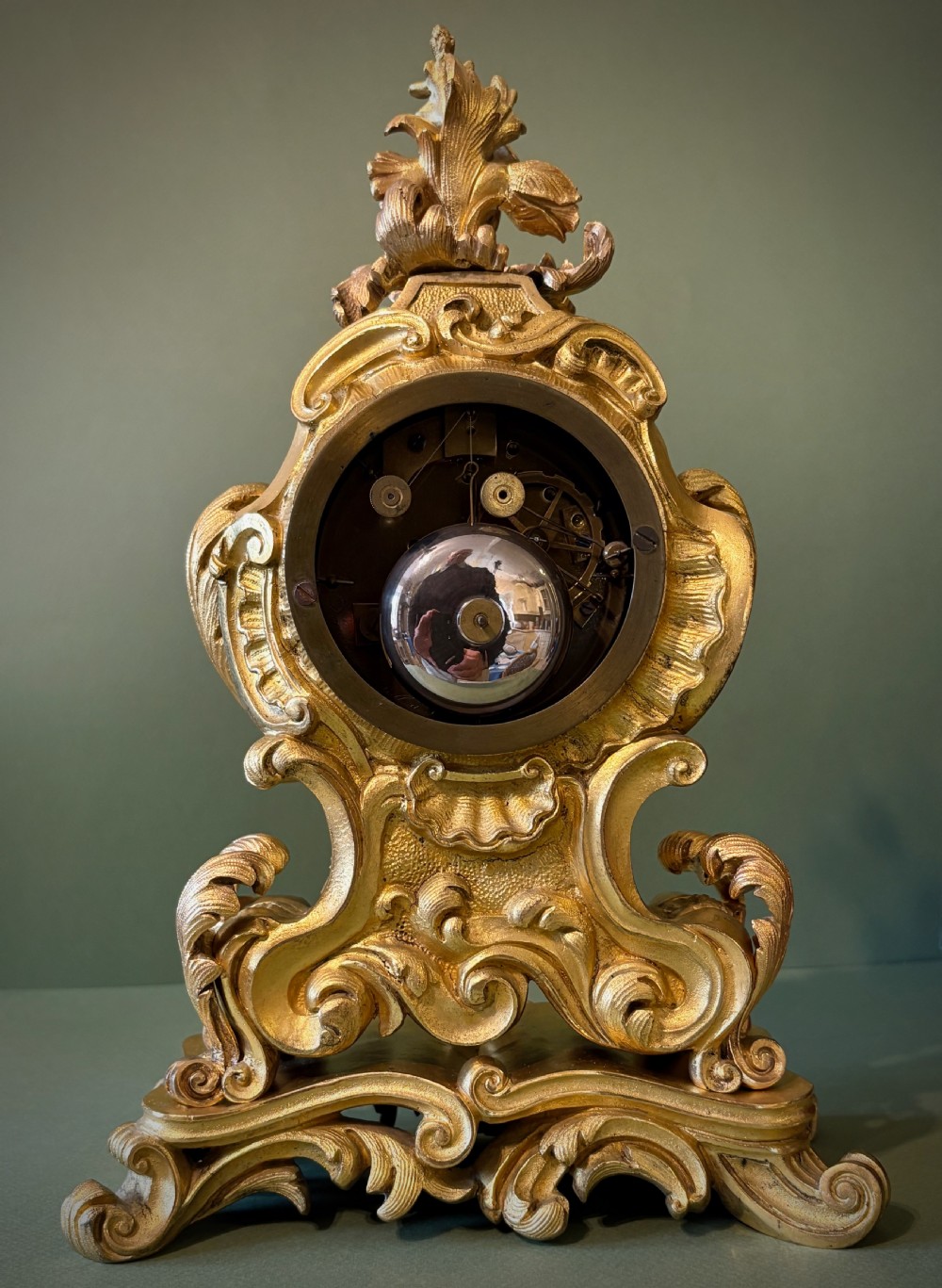 An ornate French Rococo style ormolu mantel clock featuring elaborate leaf scrollwork, a floral finial, and a circular dial with Roman numerals. View IV