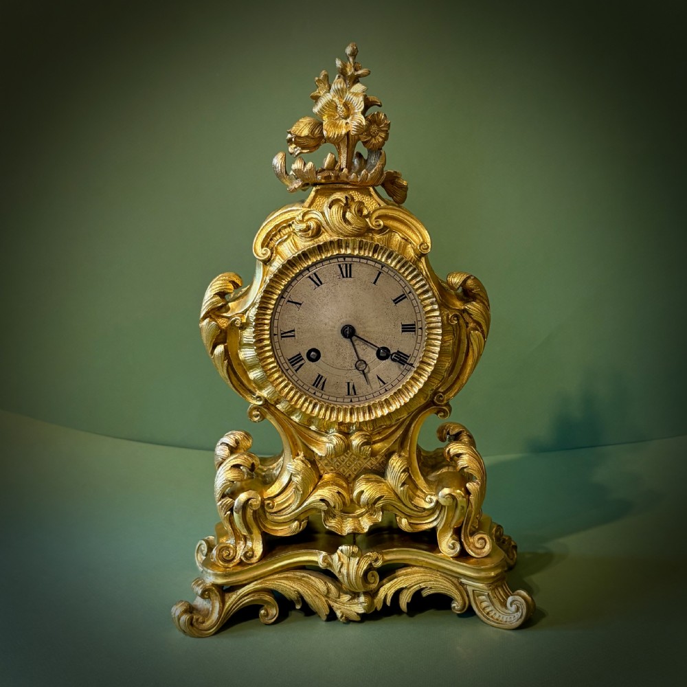 An ornate French Rococo style ormolu mantel clock featuring elaborate leaf scrollwork, a floral finial, and a circular dial with Roman numerals. View VI