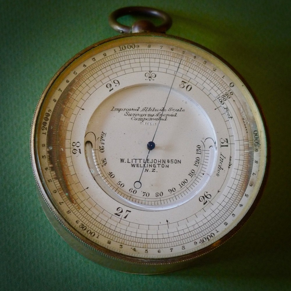A circular brass pocket aneroid barometer featuring a silvered dial with dual altitude markings and a suspension ring at the top. View III