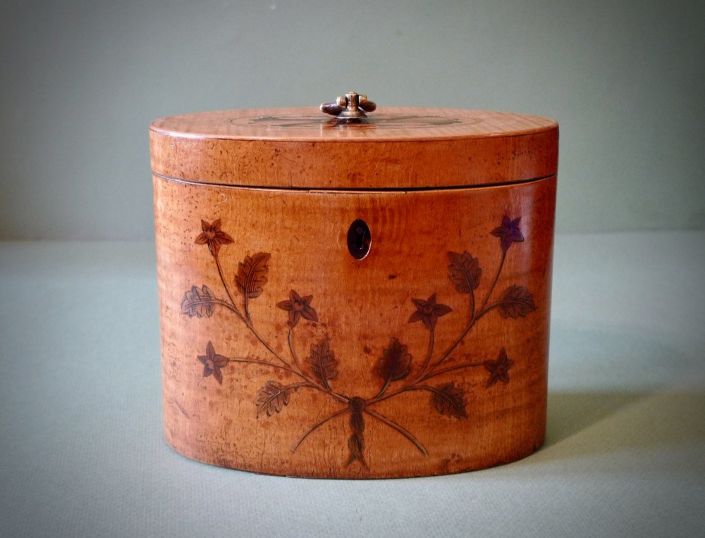 An oval Georgian satinwood tea caddy featuring floral wood inlay on the front, a top brass handle, and a central keyhole escutcheon.
