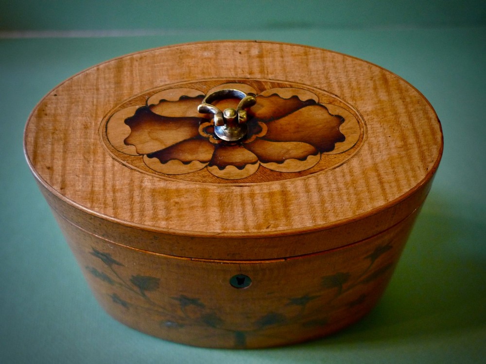 An oval Georgian satinwood tea caddy featuring floral wood inlay on the front, a top brass handle, and a central keyhole escutcheon. View II