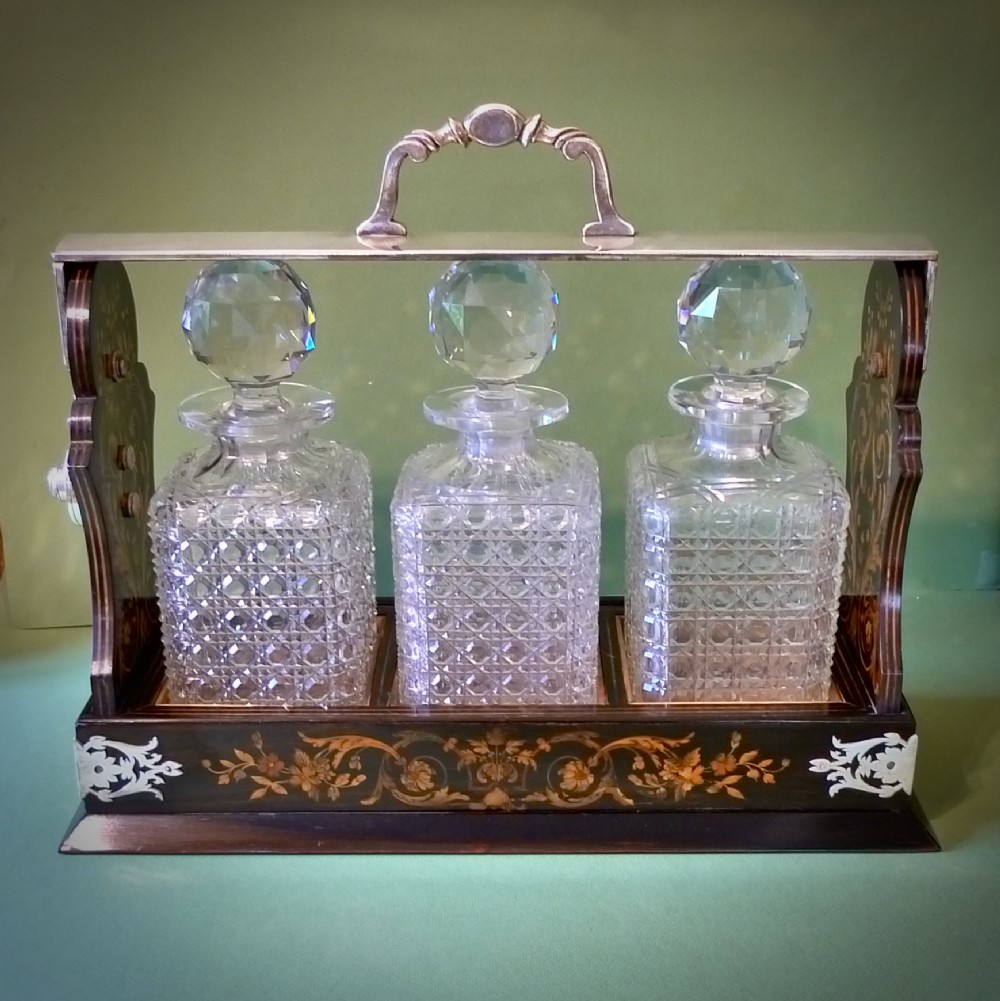 A Victorian three-bottle cut crystal decanter tantalus housed in a dark wooden frame with floral marquetry inlay and a silver-toned metal locking top handle.