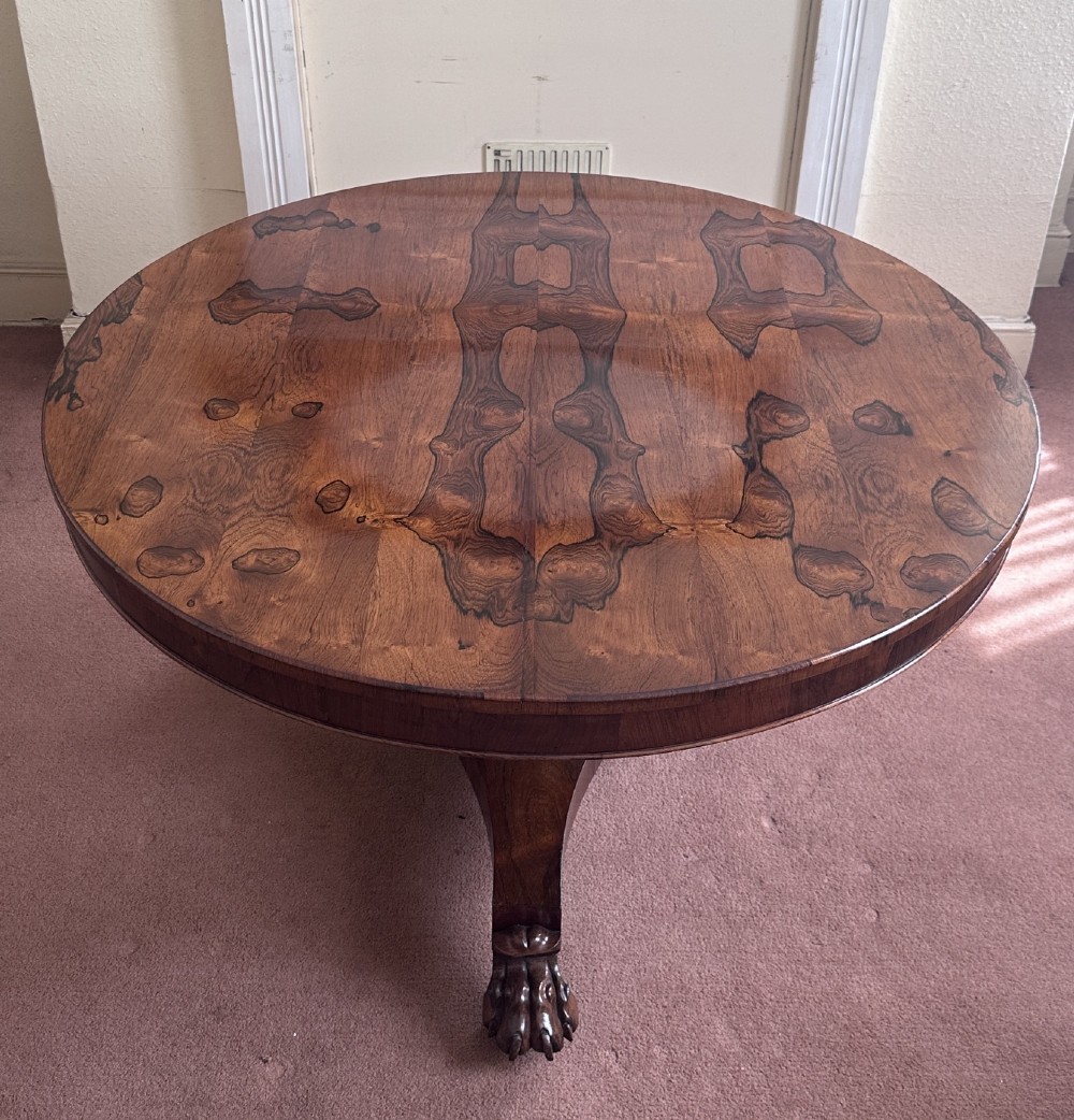 A circular William IV tilt-top rosewood centre table featuring symmetrical book-matched veneer and a central pedestal base with carved claw feet.