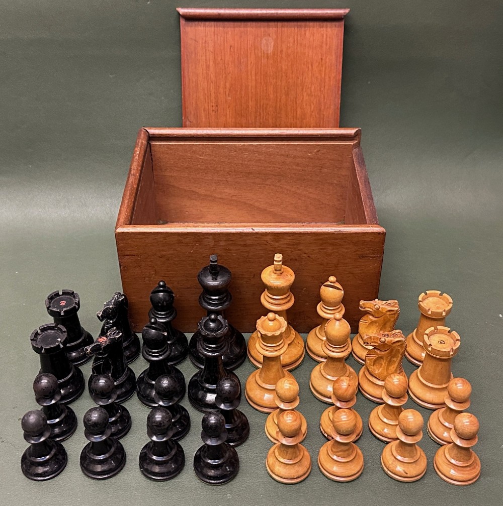 A complete set of carved boxwood and ebonised black Staunton pattern chess pieces displayed in front of a warm mahogany wooden box with a sliding lid.